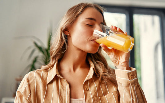 Person mit langen mittelblonden Haaren und braunem Hemd trinkt Glas Saft