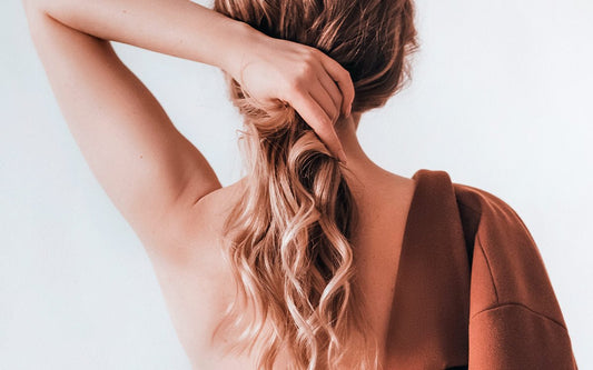 Frau von hinten, die ihre Haare zu einem Zopf hält. Das Haar ist lang, blond und leicht gewellt. 