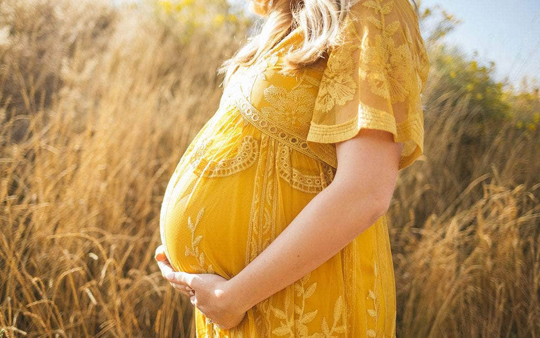Schwangere Person im gelben Kleid steht im Feld und hält ihren Bauch