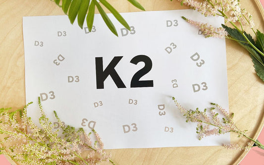 Auf einem weißen Blatt Papier ist in schwarzer und grauer Schrift K2 und D3 zu sehen. Drum herum sind grüne und weiße Pflanzen dekoriert. Das Blatt liegt auf einem runden Holzbrett.