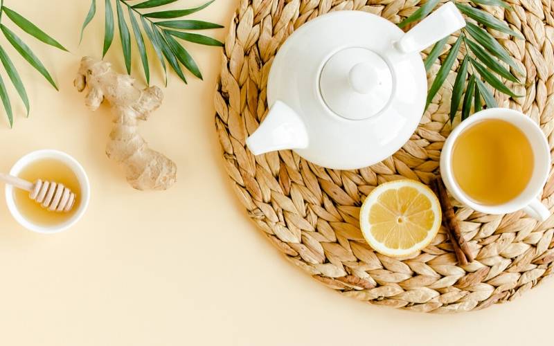 Kanne und Tasse Tee mit einer Zitronenscheibe und einer Zimtstange stehen auf einer Bastmatte auf gelbem Hintergrund