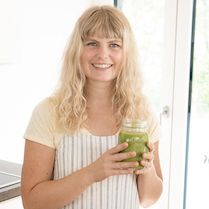 Julia steht mit grünem Smoothie an einem Tisch und lächelt 