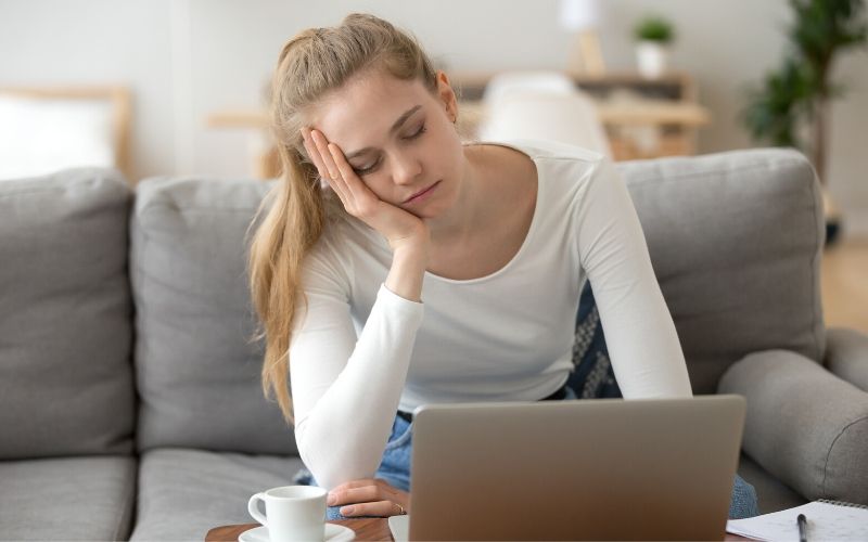 Blonde erschöpfte Frau mit fast geschlossenen Augen auf der Couch vor dem Laptop sitzen