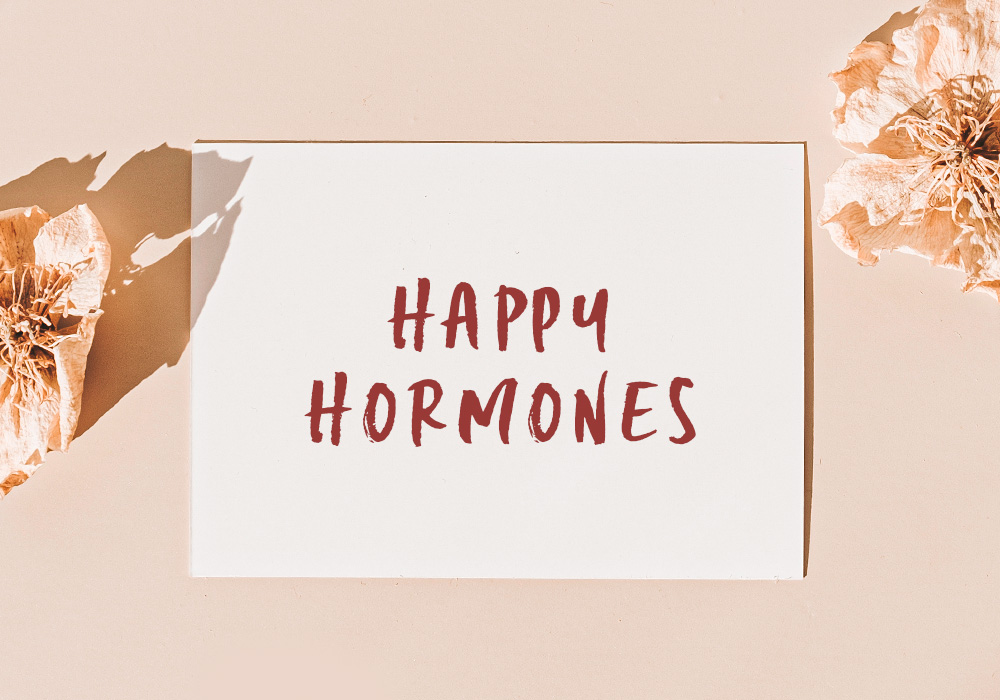 Die Worte "Happy Hormones" in weinrot auf weißem Papier und mit 2 rosa Dekoblumen