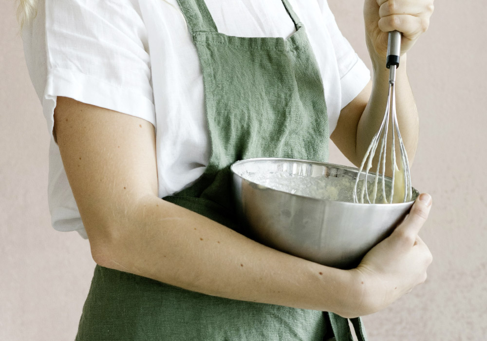 Frau mit grüner Schürze rührt mit einem Schneebesen Teig in einer großen silbernen Schüssel
