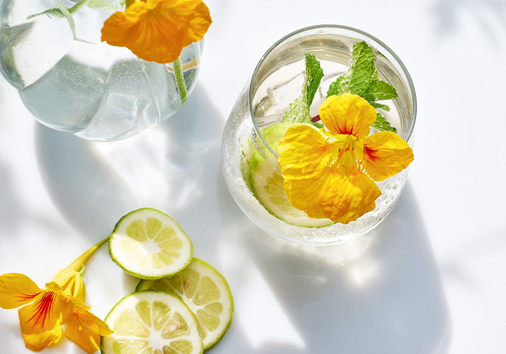 Ein frischer Sommerdrink in einem Glas. Angerichtet mit essbaren Blüten, Zitrone und Eiswürfeln.