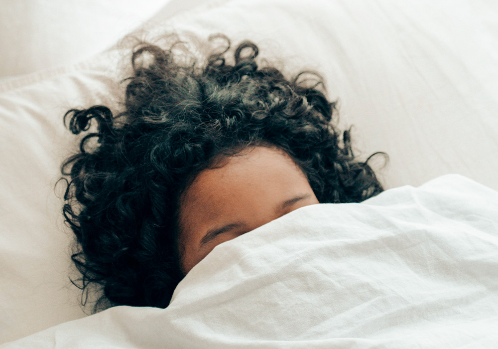 Schwarzhaarige Frau, die im Bett liegt und ihr Gesicht ist zur Hälfte von einer weißen Decke bedeckt. 