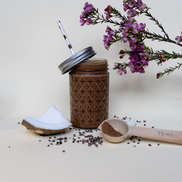 Ein Glas Rise&Shine Superfood Mix mit lila Blumen und einem Stück Kokosnuss