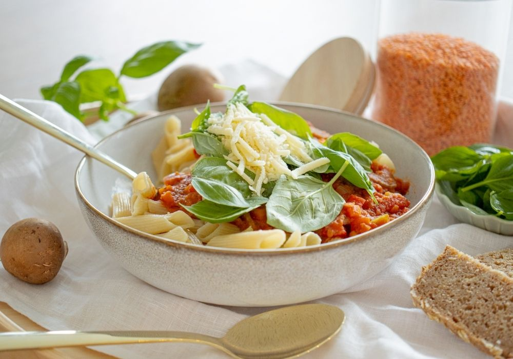 Nudeln mit Roten-Linsen-Bolognese in einer Porzellan Schüssel. Als Topping dienen Spinatblätter und veganer Käse.