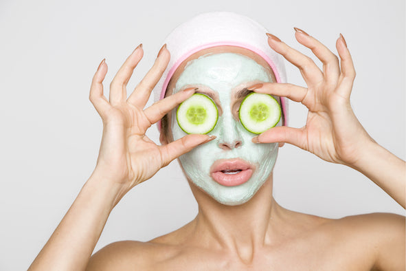 Frau mit einer Gesichtsmaske hält sich 2 Scheiben Gurken vor die Augen